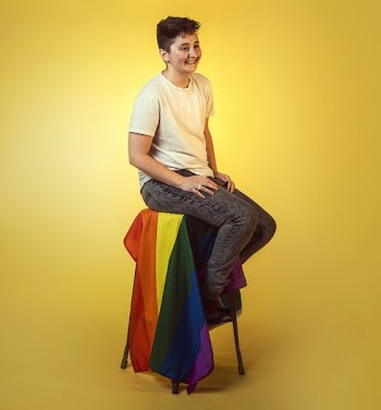 Smiling Trans Man Seated Above Draped Rainbow Flag