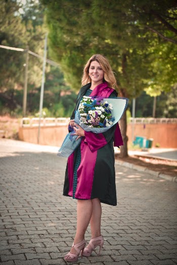 Smiling Woman in Black and Pink Academic Dress Posing on Concrete Pavement Holding a Bouquet of Flowers