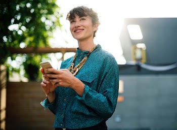 smiling woman in blue dress shirt holding gold iPhone 6 standing in front of house near green leafed tree during daytime