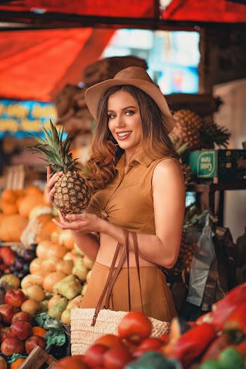 Smiling Woman Standing Beside Assorted Fruits While Holding Pineapple Fruit