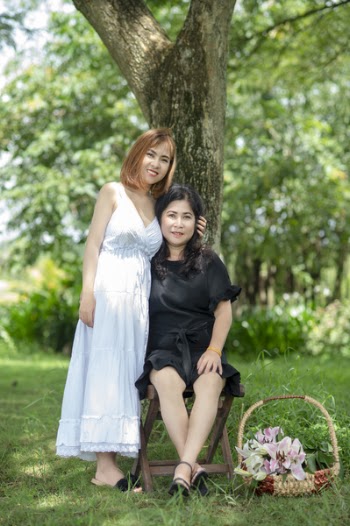 Smiling Woman Standing Beside Smiling Woman Sitting on Folding Stool Under Tree