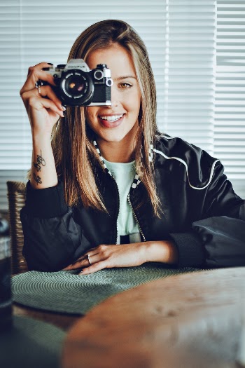 Smiling Woman Using Camera While Sitting Inside Room Beside Window Blinds