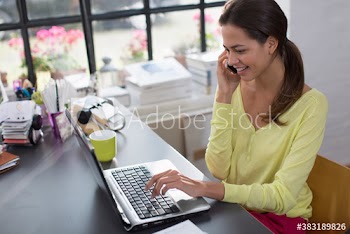 Smiling woman working at laptop and talking on smart phone in office