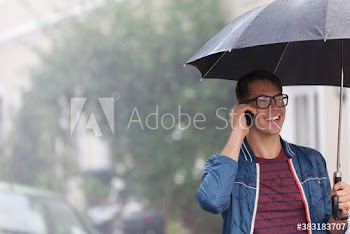 Smiling young man with umbrella talking on smart phone in rain