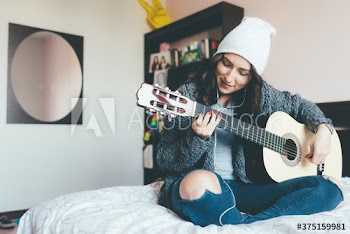 Smiling young woman playing guitar while sitting on bed at home