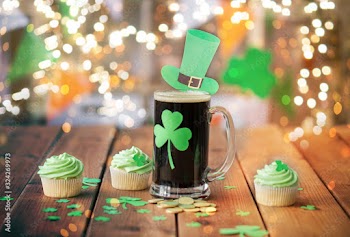 st patricks day, holidays and celebration concept - glass of dark draft beer with shamrock, green cupcakes and gold coins on wooden table over festive lights
