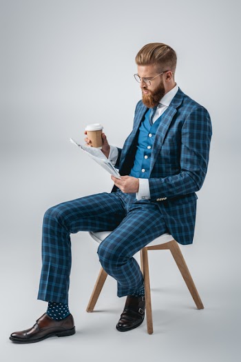 stylish man in suit with coffee to go reading newspaper while sitting on chair isolated on grey