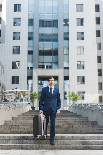 Stylish young businessman with luggage walking by business district