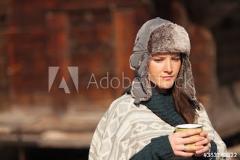 Thoughtful woman in fur hat drinking coffee outside cabin