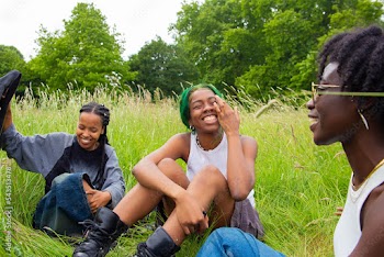 Three black non binary friend sat in the park laughing and talking.