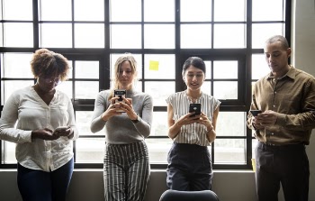 Three Women and One Man Standing While Using Phones