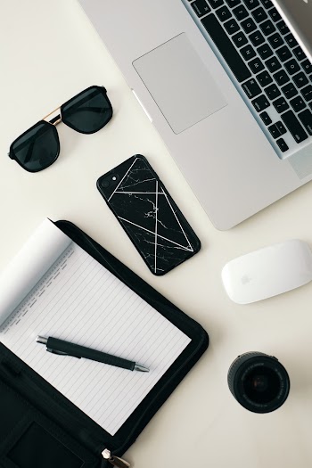 Top View Of A Pen On A Notepad, Sunglasses And Various Electronic Gadgets On A White Table Top