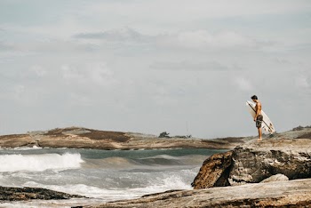 Topless Man Carrying Surfboard on Brown Rock