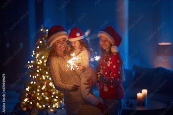 Two little sisters hugging pretty mother and smiling cheerfully while standing near christmas tree with sparklers in hands. Concept of family, winter holidays and happiness. 
