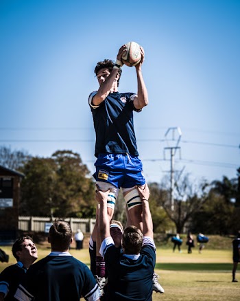 Two Men Carrying Standing Man Holding Rugby Ball over Field With People Across Electric Post Under Blue Sky