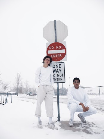 Two Men in White Sweaters Standing and Crouching Beside Road Sign