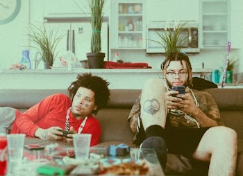 two men lying and sitting on brown sofa while holding game controller and using smartphone