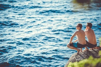 two men sitting on cliff in front of body of water
