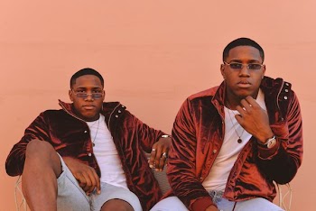 two men wearing machine clothes sitting down against pink wall