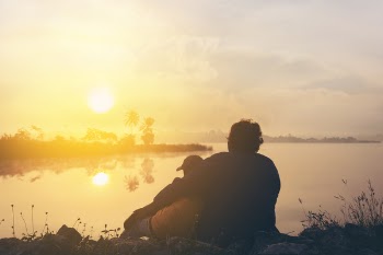 Two People On Grass Beside Body Of Water During Golden Hour