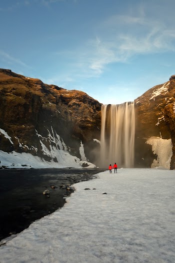 Two People Standing on Snow Facing the Waterfalls during Day
