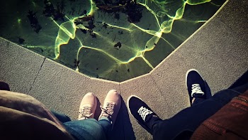 Two Person Standing Near Body of Water Taking Picture of Shoes