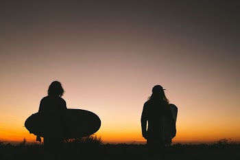 two person walking on road while holding surfboard during golden hour