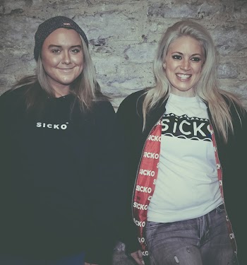 Two Smiling Women Wearing Black Jacket Leaning on Gray Wall