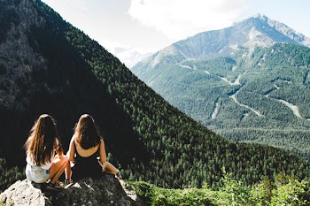 Two Women Sitting on Rock Facing Mountains Under White Cloudy Skies