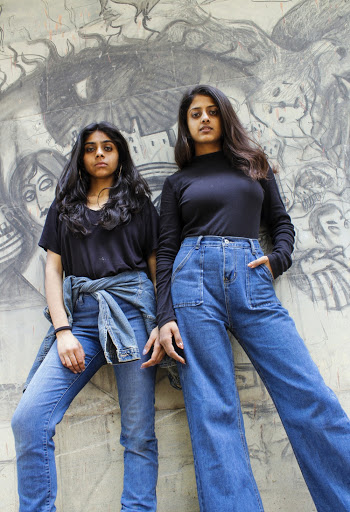 Two Women Wearing Black Tops And Blue Jeans Leaning on Concrete Wall