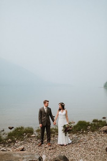 wedding couple standing beside body of water near mountain under white sky during daytime