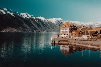 White and Brown Concrete Building Beside Body of Water Near Snow Capped Rocky Mountain Under Clear Blue Sky