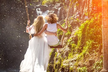 Woman and Woman Riding Wooden Swing in Front of Rock Formation