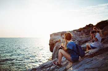 Woman holding a camera poses for a female photographer while sitting on a rocky coastline