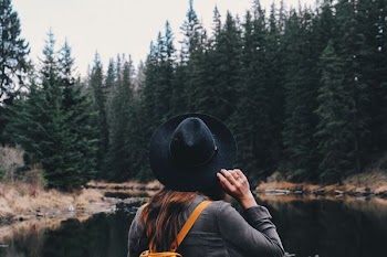 woman holding her hat while standing in front of river beside forests