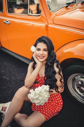 Woman  In A Polka Dot Dress Holding A Bouquet of Flowers While Sitting Down Beside An Orange Vintage Volkswagon Beetle