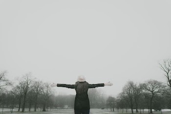 woman in black long-sleeved shirt standing and facing trees with stretched arms under white sky