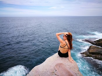 Woman in Black Swimwear Set Sitting on Top of Gray Rocky Cliff Facing Body of Water