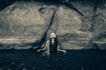 Woman in Blindfold Wearing Black Top on Body of Water While Leaning on a Rock