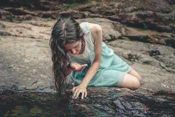 Woman in Blue and White Sleeveless Dress Sitting Beside Body of Water While Drinking Water