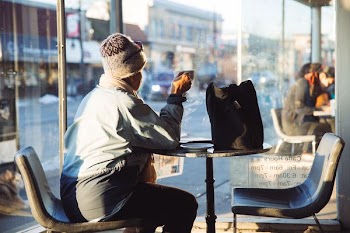 woman in blue coat and beige cap sitting at a tables with a cup of coffee at near glass walls at daytime