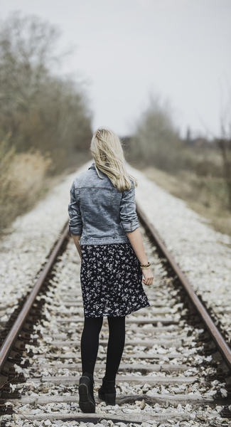 Woman in Blue Denim Jacket, Black Leggings and Black and White Dress Walking on Train Rail