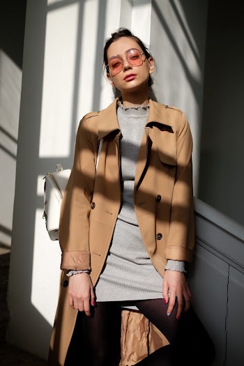 Woman in Brown Button-up Coat and Gray Top Leaning on White Wall