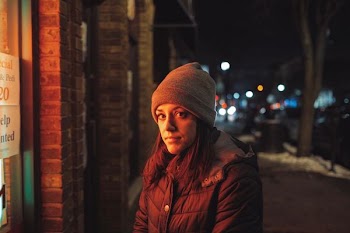 woman in brown hooded coat and brown beanie standing beside a brown building at night
