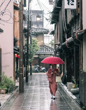 Woman in Brown Robe Holding Umbrella Walking on Concrete Pathway