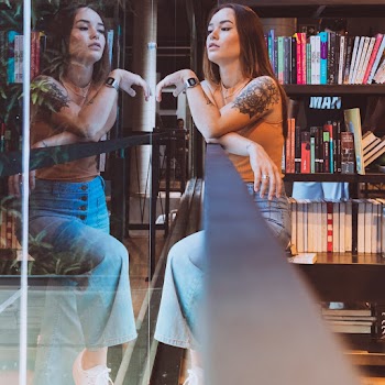 Woman in Brown Tank Top and Blue Denim Jeans Standing Near Glass Wall