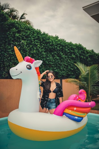 Woman In Denim Shorts And A Open Buttoned Shirt Over A Bikini Top Kneeling on White Inflatable Unicorn