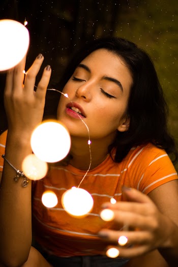 Woman in Orange T-shirt With Her Eyes Closed Holding Lit String Lights