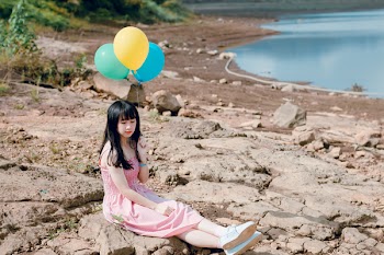 Woman in Pink Thick Strap Dress Holding a Yellow and Blue Balloons While Sitting on Rock Terrain