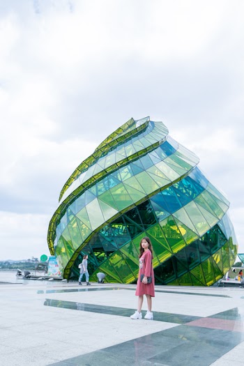 Woman in Red Dress Standing in Front of Green Glass Elliptical Building
